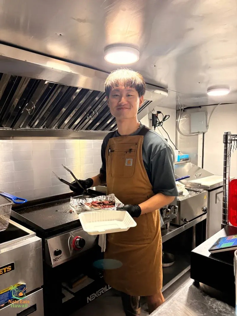 Rice & Friesのオーナーシェフ・David。茶色いCarharttのエプロンを着て、トングと料理の容器を持ちながらフードトラックのキッチンで調理中
