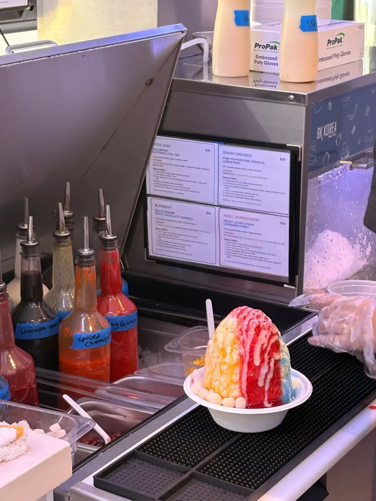 Sin Ice Stir housemade syrups in bottles with shave ice being prepared behind the counter